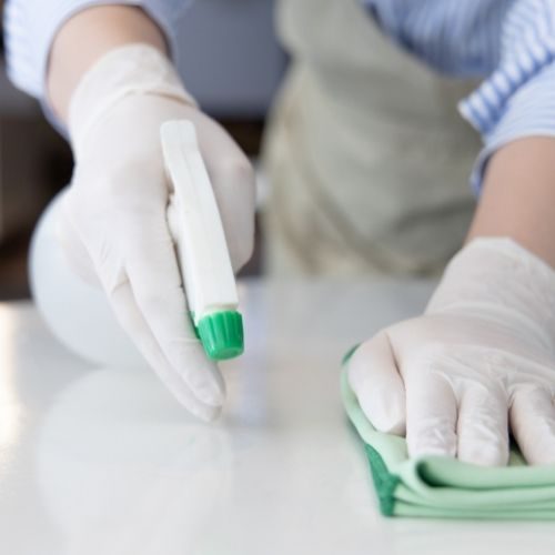 A hand wearing white rubber gloves spraying cleaning solution onto a countertop and using a sponge to wipe it off.