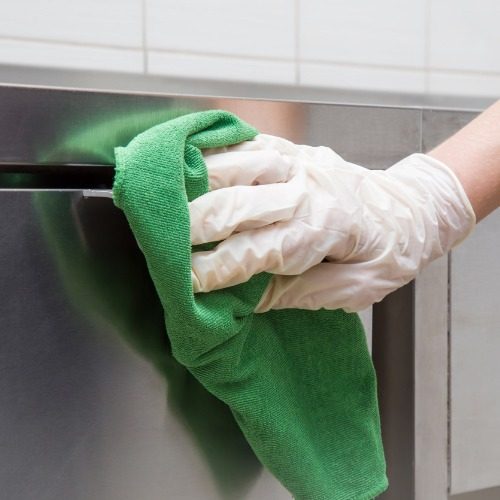 A hand wearing a white rubber glove wiping the front of a stainless steel dishwasher with a green microfiber cleaning cloth.